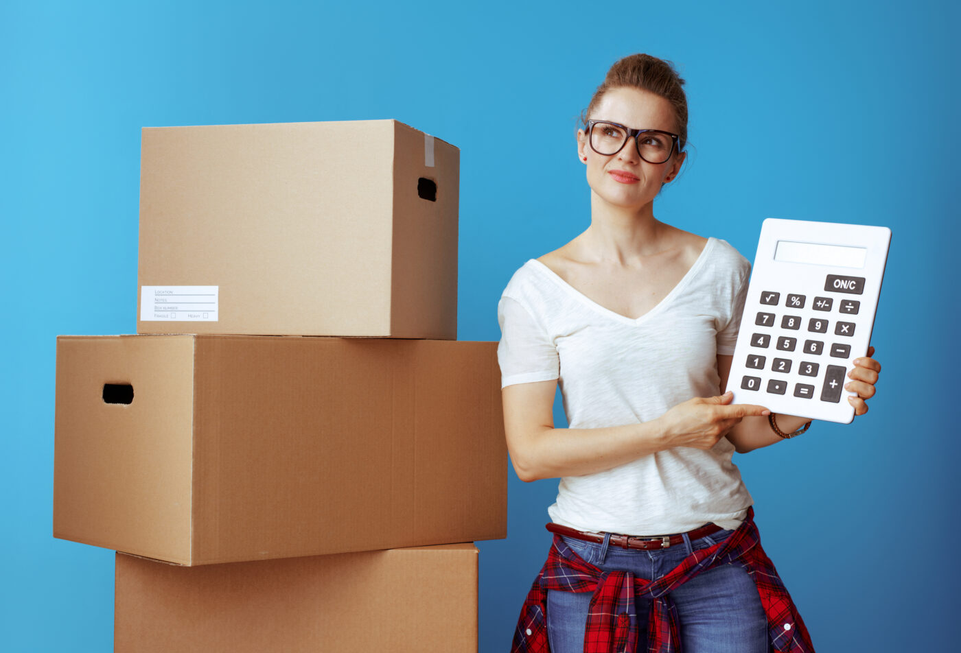 Portrait of pensive modern woman in white t-shirt near cardboard box with calculator against blue background. stay on budget.