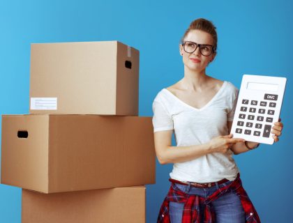 Portrait of pensive modern woman in white t-shirt near cardboard box with calculator against blue background. stay on budget.